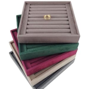 May include: A stack of five rectangular jewelry display trays with velvet-like lining in taupe, dark green, burgundy, beige, and gray. Each tray features multiple horizontal slots for rings. A single gold-toned multi-band ring rests on the top tray.