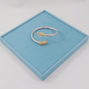 May include: A bypass bracelet featuring a twisted silver-tone cable band that terminates in two gold-tone dragon heads. Each dragon head is detailed with small, clear pavé-set stones along the top, creating a textured and metallic appearance.