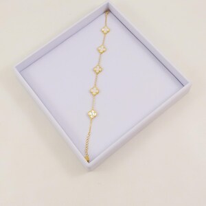 Puede incluir: Una pulsera de cadena dorada con cinco dijes blancos en forma de trébol de cuatro hojas, espaciados uniformemente y con bordes dorados. La pulsera incluye una cadena de eslabones finos y una sección de extensión ajustable, presentada en una caja.