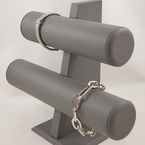 May include: A two-tier grey jewellery display stand featuring a matte finish. The top bar holds a silver mesh bracelet, while the bottom bar displays a silver chain-link bracelet with large, flat, rectangular links and a polished metallic finish.