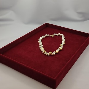 May include: A pearl necklace with gold accents rests on a red velvet tray. The necklace features white pearls interspersed with gold-colored beads. The tray is rectangular and has a deep red color.