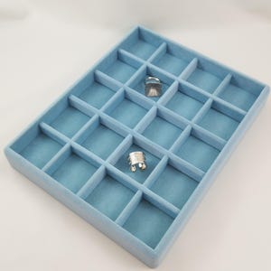 May include: A light blue rectangular jewellery organiser tray with twenty individual compartments. The tray features a soft velvet-like texture and contains two silver-toned metal rings placed within separate sections of the grid.