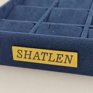 Puede incluir: Una caja de joyería de terciopelo azul con una etiqueta dorada que dice "SHATLEN".
