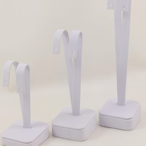 May include: Three white earring display stands of different sizes. Each stand has a square base and a curved, upright piece with two holes for earrings.