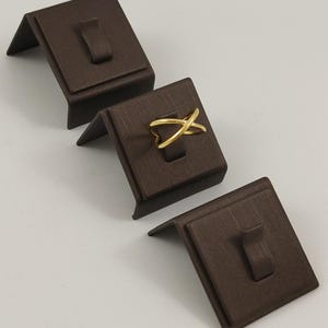 May include: A gold-toned ring featuring a crossover design, displayed on a dark brown textured stand. The ring consists of two intersecting bands that form an X shape. Two additional empty dark brown stands are positioned nearby.