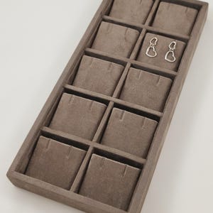May include: A rectangular, taupe-colored velvet jewelry display tray featuring ten individual compartments. Each compartment includes two small slits for securing jewelry. A pair of silver-toned heart-shaped earrings is placed in one of the sections.