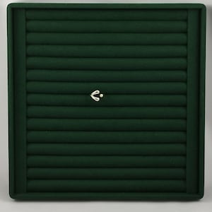 May include: A square dark green velvet jewelry display tray featuring multiple horizontal padded slots for organizing rings or earrings. A small, silver-toned metallic heart-shaped charm with a single dot accent is centered on the tray.