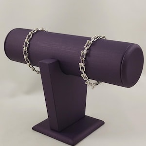 以下が含まれることがあります： 紫色の円筒形ジュエリースタンドに展示された2本の銀色の金属製チェーンブレスレット。左側のブレスレットはツイストオーバルリンクのデザインで、右側のブレスレットは小さな丸いリングで接続された長方形のU字型リンクで構成されています。