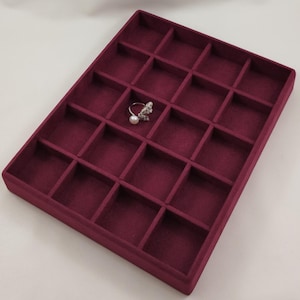 May include: A rectangular burgundy velvet jewelry display tray featuring eighteen individual square compartments. A single silver-toned ring with a white pearl and a textured, leaf-like metal design rests inside one of the center compartments.