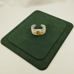 May include: A cuff bracelet featuring a central oval-shaped amber-colored stone surrounded by a border of small clear stones. The band is composed of a light cream-colored material with silver-toned edges, forming a wide, open-ended circular shape.