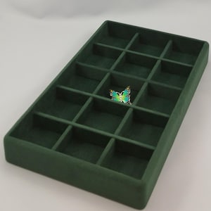 May include: A rectangular dark green velvet jewellery organiser tray featuring fifteen individual square compartments. A small, vibrant butterfly-shaped ring with blue, green, and yellow enamel wings sits inside one of the centre sections.