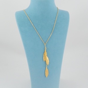 May include: A gold-colored necklace with a delicate chain and three feather pendants. The necklace is displayed on a light blue jewelry bust against a white background. The feathers have a textured surface.