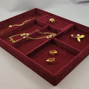 May include: A rectangular jewelry organizer tray with a deep burgundy velvet lining and multiple compartments. The tray contains a gold chain necklace, a gold ring, a gold brooch with a white pearl, and a pair of gold stud earrings.