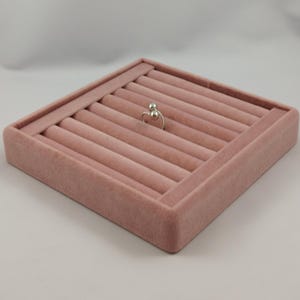 May include: A silver ring featuring two small spheres at the top, positioned in the center of a square, dusty pink velvet ring display tray with multiple horizontal slots for organizing and showcasing jewelry pieces.