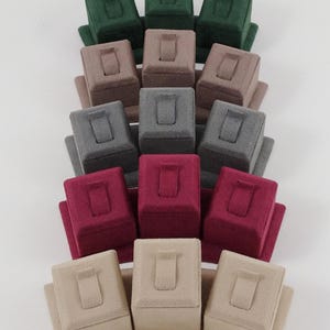 May include: A collection of fifteen velvet-covered jewelry display stands arranged in five rows of three. The stands feature a rectangular base and a raised central block with a vertical slot for rings. Colors include forest green, taupe, gray, burgundy, and beige.