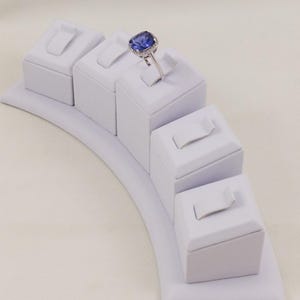 Könnte beinhalten: Weißer Ringständer mit fünf einzelnen gepolsterten Fächern zum Aufbewahren von Ringen. Der Ständer ist gebogen und hat einen weißen Hintergrund.