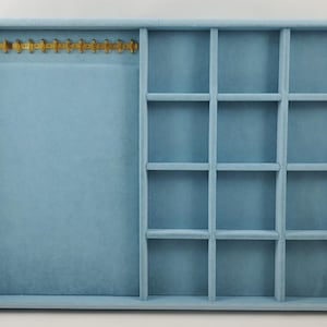 Peut inclure: Un plateau organisateur de bijoux doublé de velours bleu clair, comprenant une grande section rectangulaire ouverte sur la gauche avec une barre à collier en métal doré en haut et douze petits compartiments carrés sur la droite.
