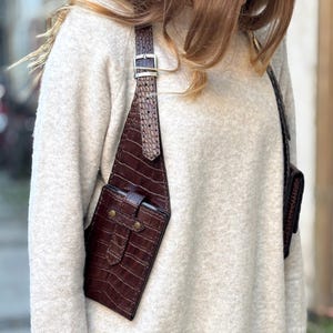 May include: A brown leather crossbody bag with a crocodile pattern. The bag has two pockets and a strap that goes over the shoulder.