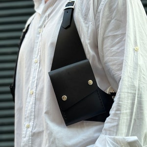 May include: A black leather chest rig with a single pocket and silver metal accents. The rig is worn over a white button-down shirt.