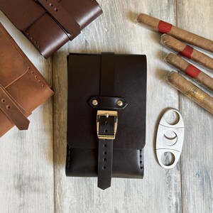 May include: A set of three leather cigar cases with a metal cigar cutter. The cases are brown and black and have a strap with a buckle. The cutter is silver and has a crescent moon shape.