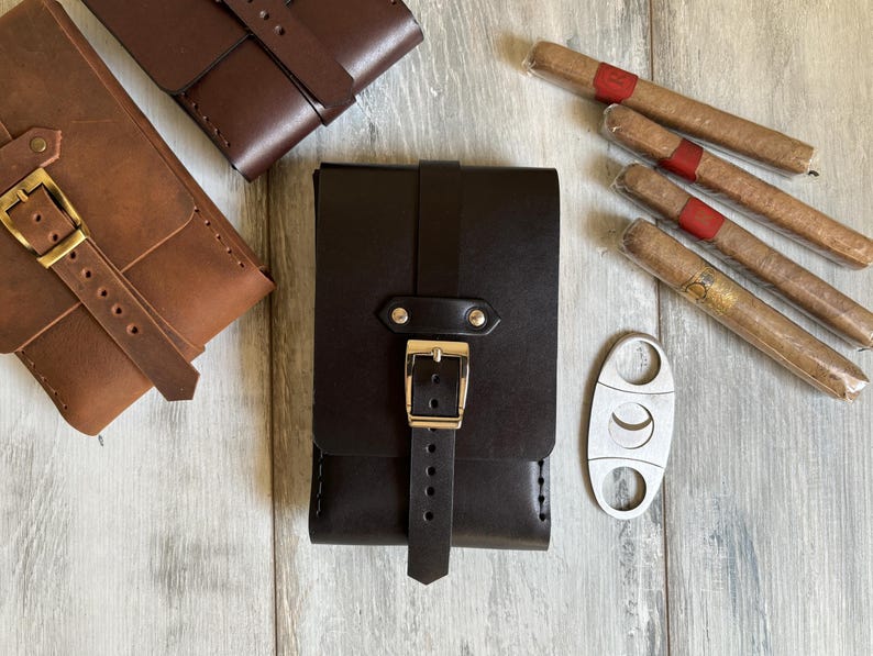 May include: A set of three leather cigar cases with a metal cigar cutter. The cases are brown and black and have a strap with a buckle. The cutter is silver and has a crescent moon shape.