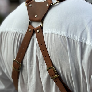 Puede incluir: Correa de c&aacute;mara de cuero marr&oacute;n con hebillas y anillos de lat&oacute;n. La correa se lleva sobre una camisa blanca.