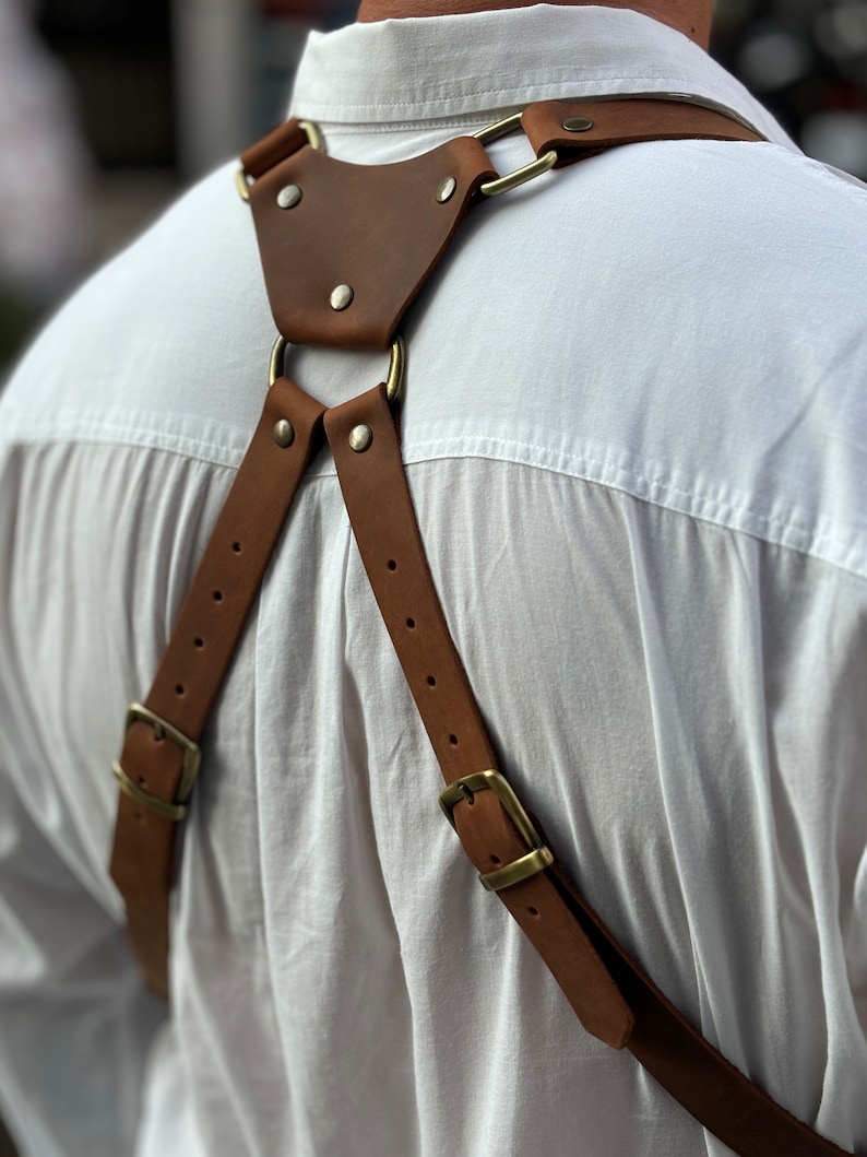 Puede incluir: Correa de c&aacute;mara de cuero marr&oacute;n con hebillas y anillos de lat&oacute;n. La correa se lleva sobre una camisa blanca.