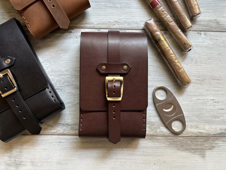 May include: Three leather cigar cases with a metal cigar cutter. The cases are brown, black, and dark brown. The cases have a buckle closure and a strap.