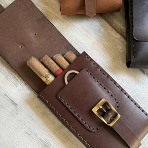 May include: A brown leather cigar case with a buckle closure and a pocket for a cigar cutter. The case has four slots for cigars and is lined with brown fabric.