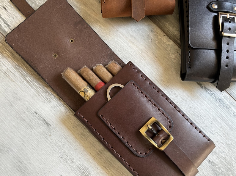 May include: A brown leather cigar case with a buckle closure and a pocket for a cigar cutter. The case has four slots for cigars and is lined with brown fabric.