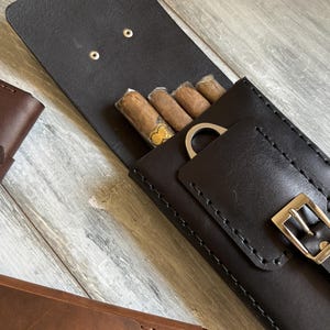 May include: A black leather cigar case with a built-in cutter and a buckle closure. The case is open and four cigars are visible inside.