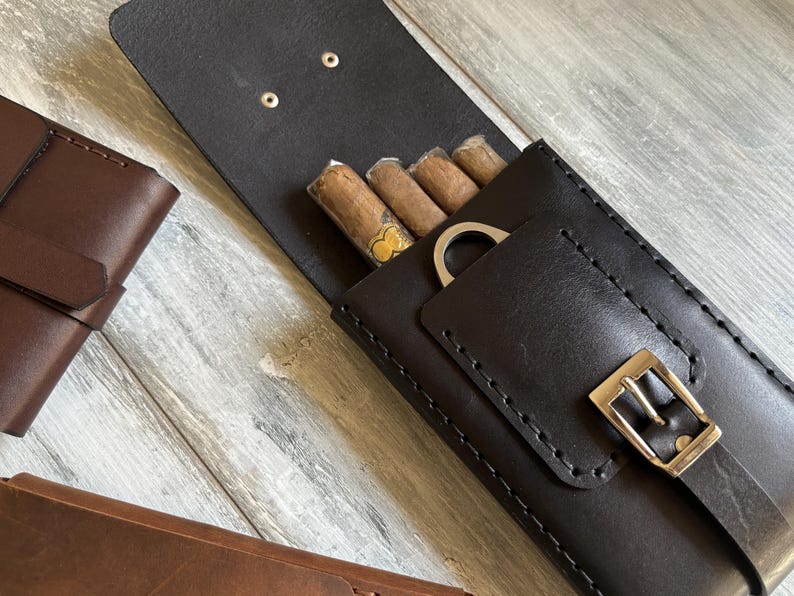 May include: A black leather cigar case with a built-in cutter and a buckle closure. The case is open and four cigars are visible inside.