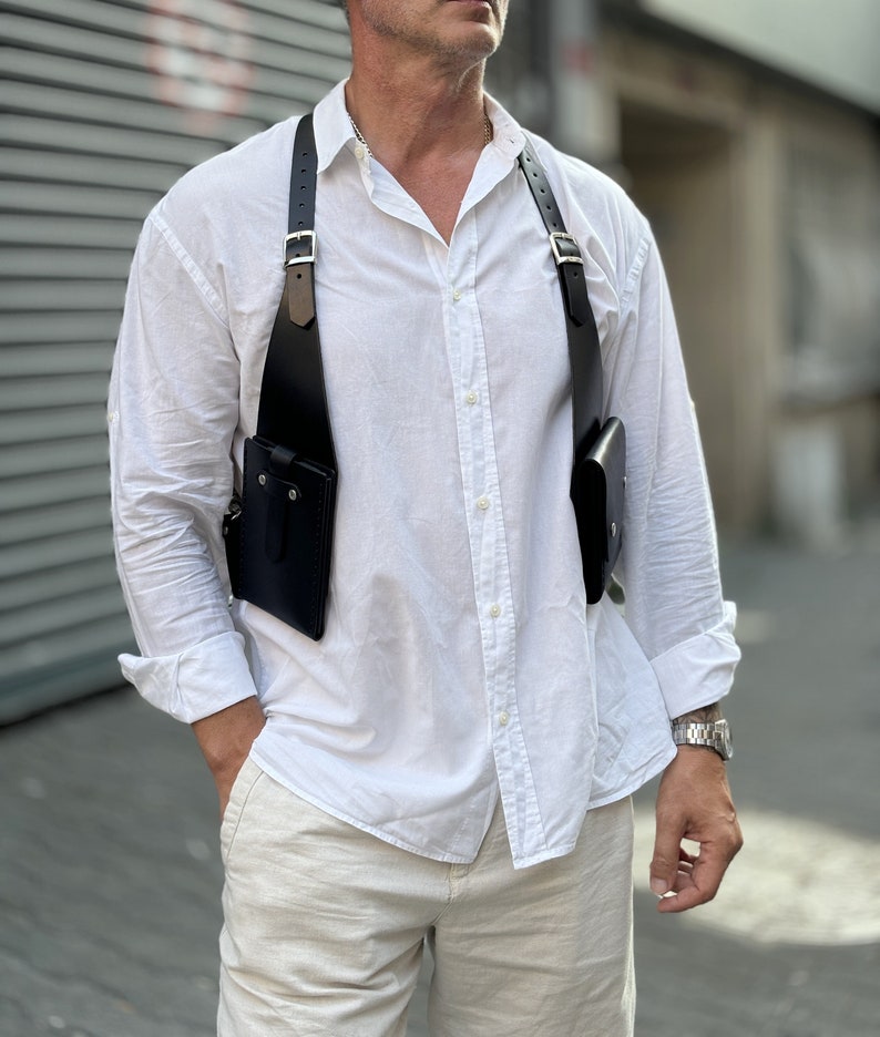 May include: A black leather chest rig with two pockets and adjustable straps. The rig is worn over a white button-down shirt and beige trousers.