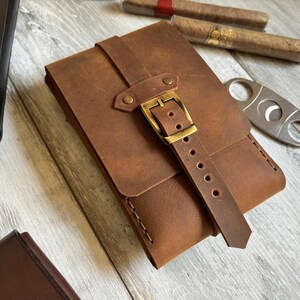 May include: A brown leather cigar case with a brass buckle and strap. The case has a flap that closes with a snap.
