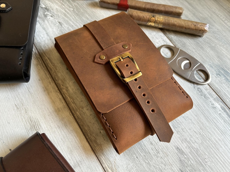 May include: A brown leather cigar case with a brass buckle and strap. The case has a flap that closes with a snap.