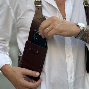 Puede incluir: Una funda de tel&eacute;fono de cuero marr&oacute;n con una correa que se coloca sobre el hombro. La funda tiene una solapa que cubre el tel&eacute;fono y una hebilla de metal. Se puede ver un tel&eacute;fono inteligente dentro de la funda.