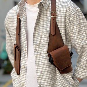 Peut inclure: Un sac à bandoulière en cuir marron avec une fermeture à rabat et une boucle. Le sac a une bandoulière qui passe par-dessus l'épaule et traverse la poitrine. Le sac est porté par une personne portant une chemise blanche à carreaux.