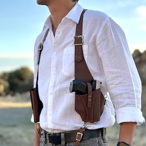 Op de afbeelding: Een bruine leren schouderholster met een pistool erin. Het holster heeft een band die over de schouder gaat en een gesp aan de zijkant. Het holster wordt gedragen door iemand die een wit shirt en jeans draagt.