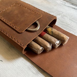 May include: A brown leather cigar case with a flap that opens to reveal a compartment for cigars. The case has a metal cigar cutter attached to the flap.