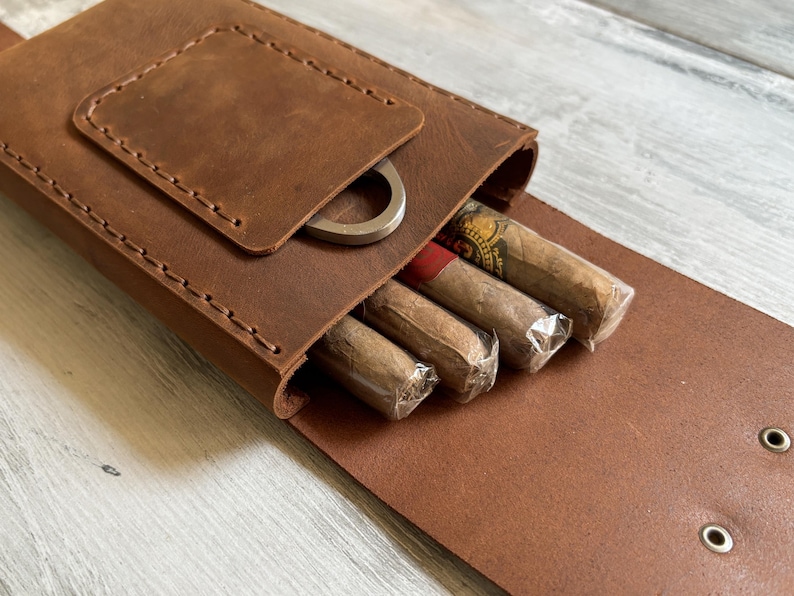 May include: A brown leather cigar case with a flap that opens to reveal a compartment for cigars. The case has a metal cigar cutter attached to the flap.