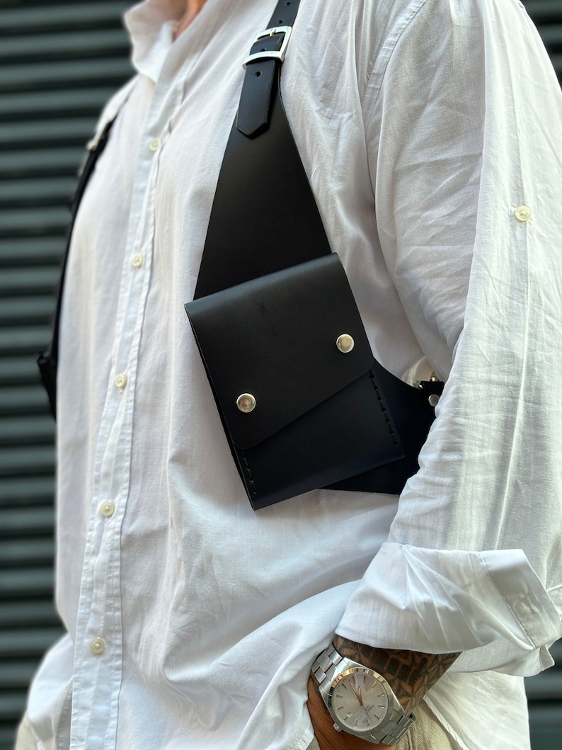 May include: A black leather chest rig with a single pocket and silver metal accents. The rig is worn over a white button-down shirt.