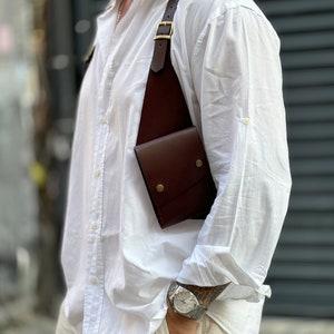 May include: A brown leather crossbody bag with a strap that goes over the shoulder. The bag has a flap closure and a small pocket on the front. The bag is worn by a person wearing a white shirt and beige trousers.