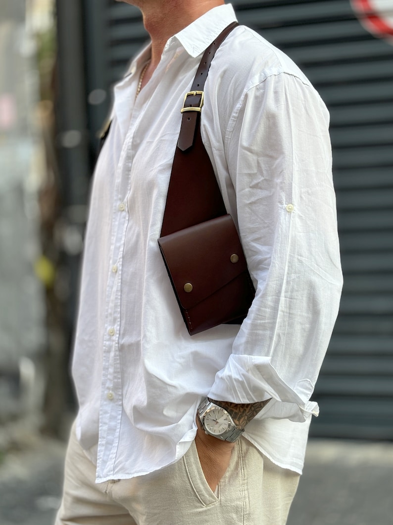 May include: A brown leather crossbody bag with a strap that goes over the shoulder. The bag has a flap closure and a small pocket on the front. The bag is worn by a person wearing a white shirt and beige trousers.