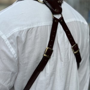 May include: A brown leather camera strap with brass hardware. The strap is worn over the shoulders and has a criss-cross design at the back.