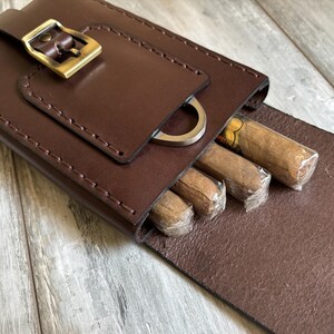 May include: A brown leather cigar case with a metal buckle and a cigar cutter. The case is open and contains several cigars.