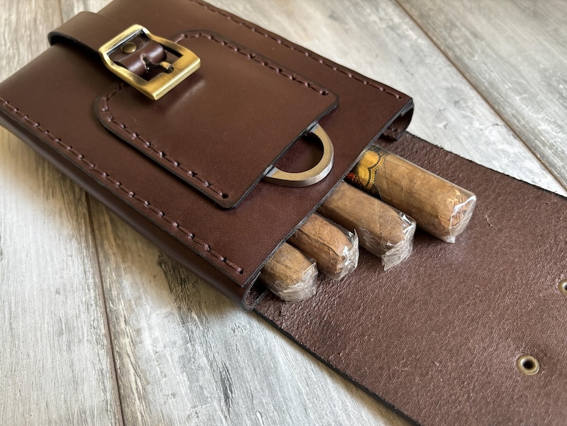 May include: A brown leather cigar case with a metal buckle and a cigar cutter. The case is open and contains several cigars.