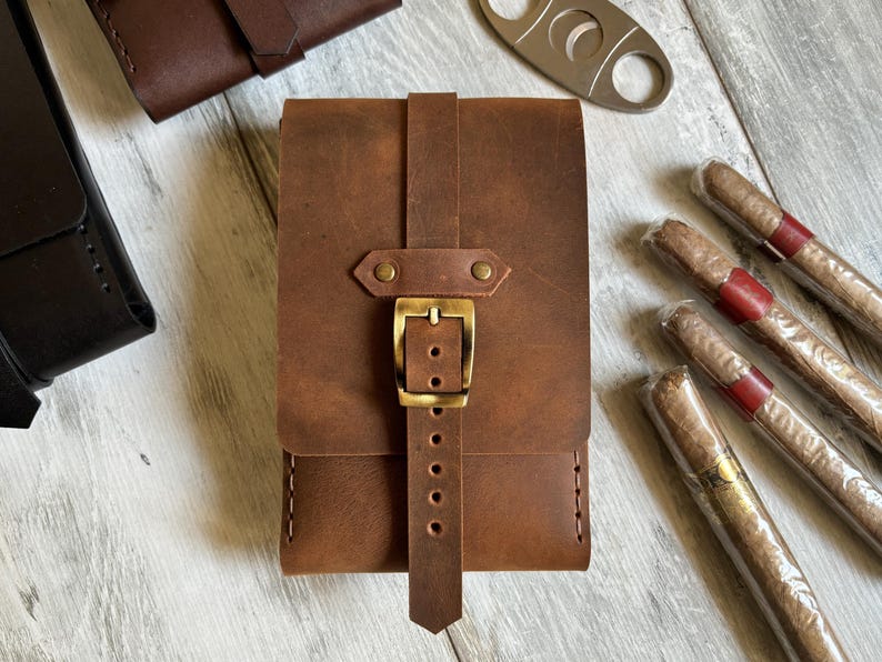 May include: A brown leather cigar case with a brass buckle and stitching. The case has a flap that closes with a strap.