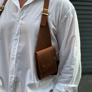 Puede incluir: Un bolso de cuero marr&oacute;n cruzado con un solo bolsillo y una correa con hebilla. El bolso se lleva sobre una camisa blanca de botones.