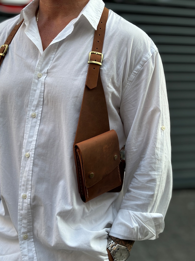 Puede incluir: Un bolso de cuero marr&oacute;n cruzado con un solo bolsillo y una correa con hebilla. El bolso se lleva sobre una camisa blanca de botones.