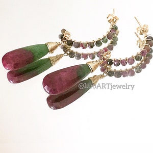 May include: Gold hoop earrings with a double row of pink and green gemstones. The earrings have teardrop-shaped pink and green gemstones dangling from the bottom.